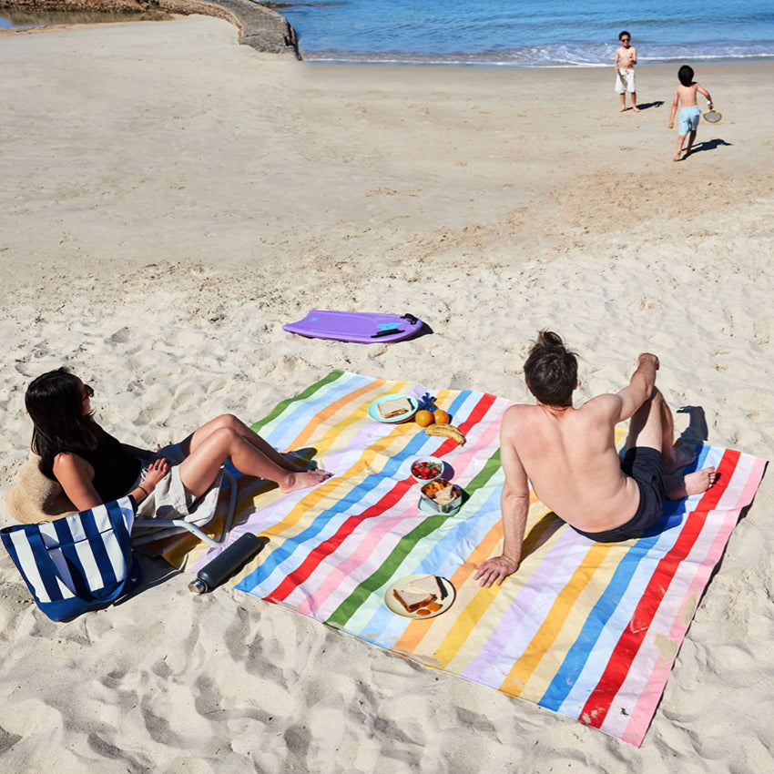 The Dock & Bay Picnic Blanket in Candy Stripe laid out on a beach with two people sitting on it