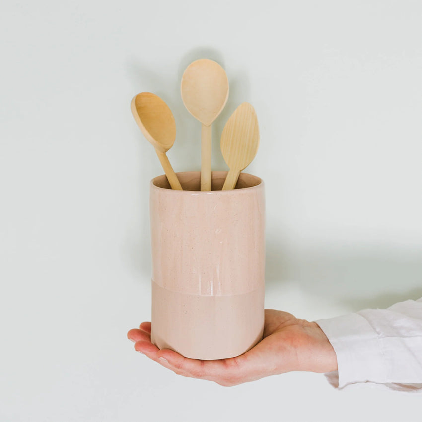 The JS Ceramics Utensil Holder in Pink Sand on someones hand with wooden spoons on it on a blank background