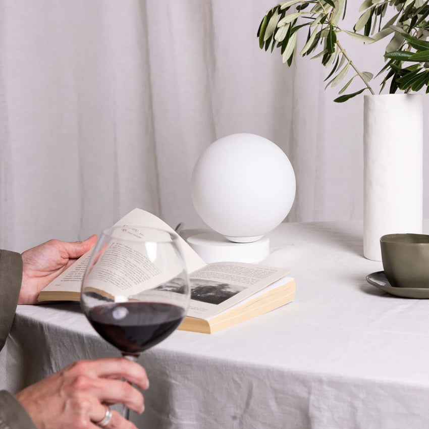 The Ned Collections Yuri Table Lamp turned off on a table with a vase, book and person having a glass of wine