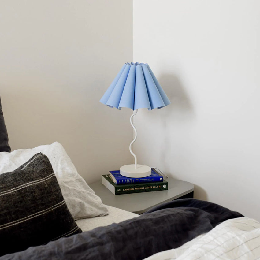 The Paola and Joy Cora Table Lamp in Tranquil Blue next to a bed on a bedside table and books