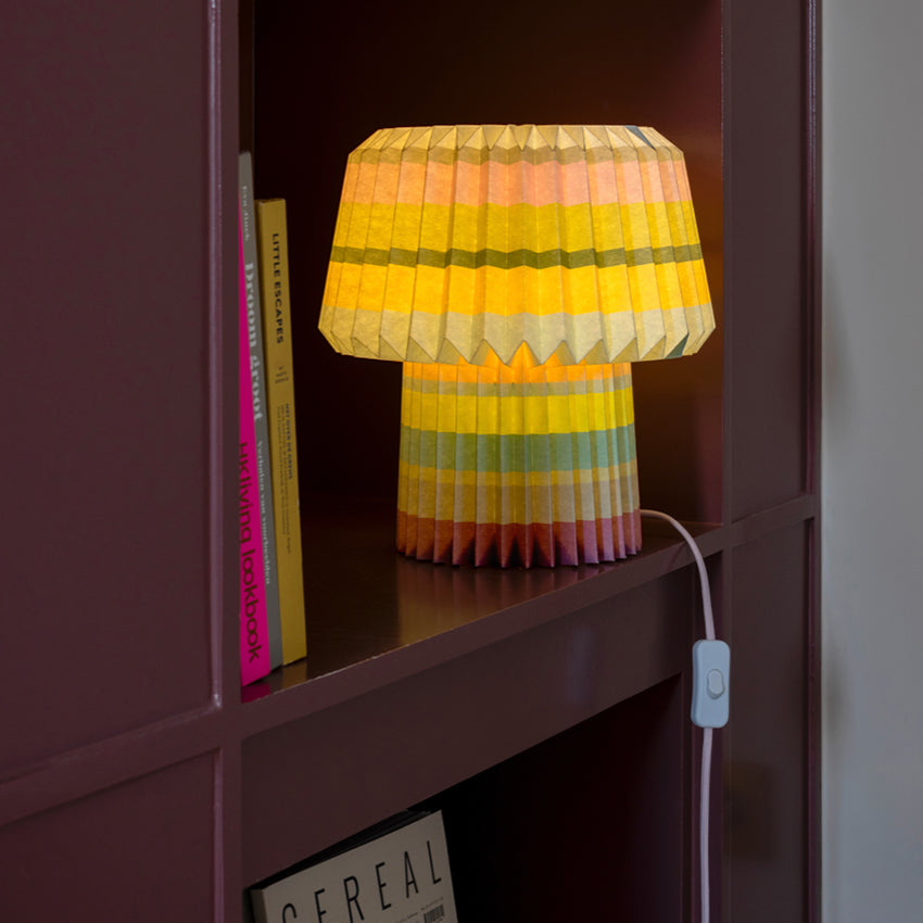 The Remember Paper Lamp in Anna Stripes print glowing on a maroon bookshelf