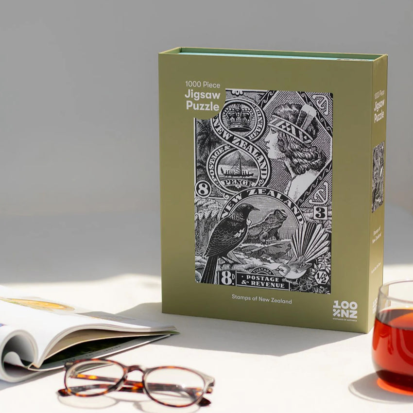 The 100 Percent NZ Stamps of NZ Jigsaw Puzzle on a table with a magazine, glasses and a drink