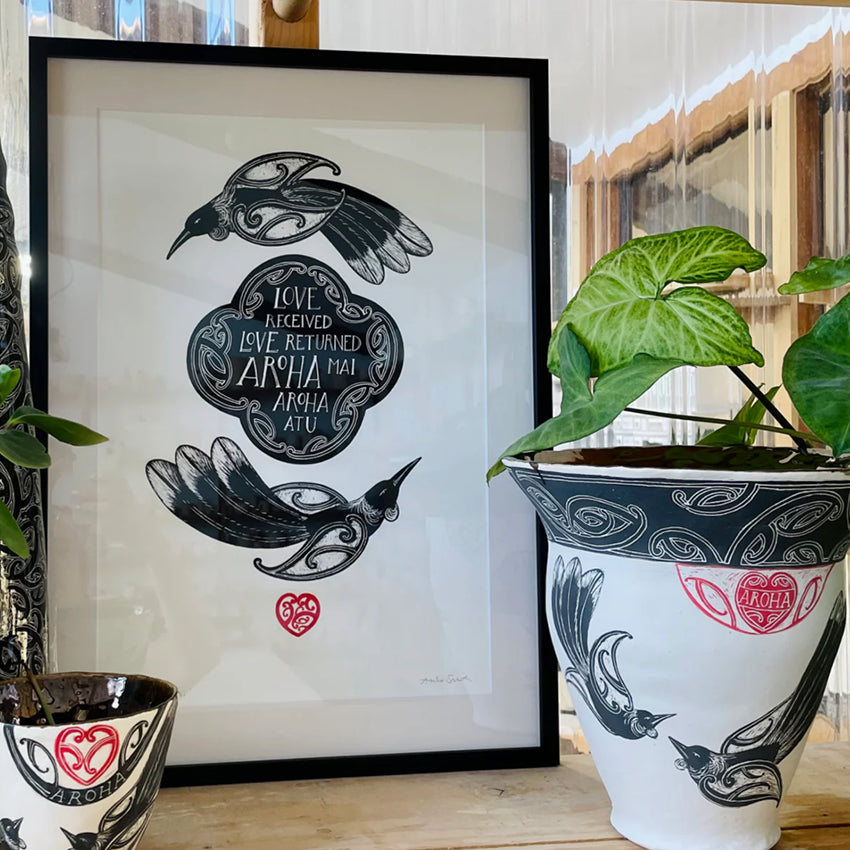 The Amber Smith Aroha Mai Art Print in a black frame next to a vase