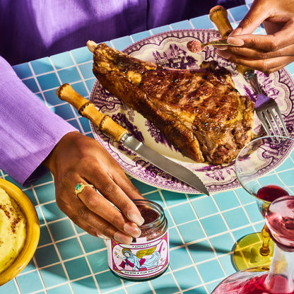 The Apostle Red Wine and Plum Herb Salt Seasoning jar being placed on a tiled table next to a plate of meat