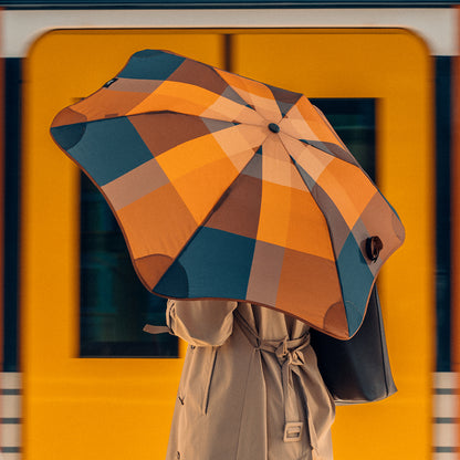 The Blunt Metro Umbrella in Limited Edition Gingerbread with model holding it in front of a train