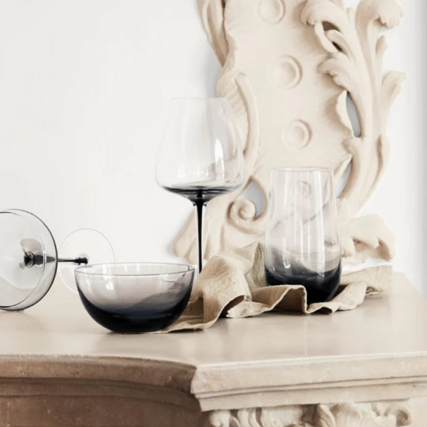 The Broste Copenhagen Tall Tumbler Glass in Smoke on a mantel with other glassware