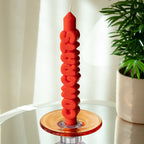 The Candle Hand Congrats 3D Taper Candle in orange in a glass candle holder next to a plant