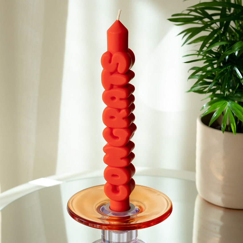 The Candle Hand Congrats 3D Taper Candle in orange in a glass candle holder next to a plant