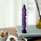 The CandleHand Happy Bday Taper Candle in purple in a candle holder on top of books next to walnuts