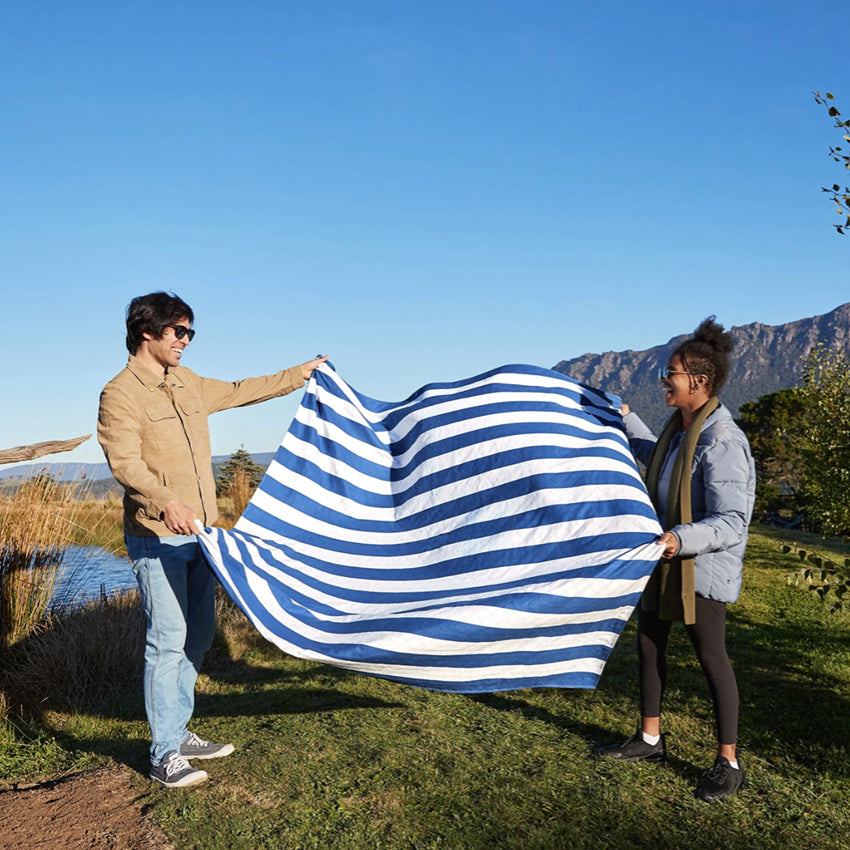 Two people shaking out the Dock & Bay Picnic Blanket in Whitsunday Blue and White stripe