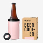The Huski Beer Cooler in Powder Pink with a beer bottle in it next to its box on a blank background