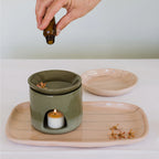 The JS Ceramics Ceramic Oil Burner in Sage Green on a platter with a candle in the bottom, essential oil being dropped in the top