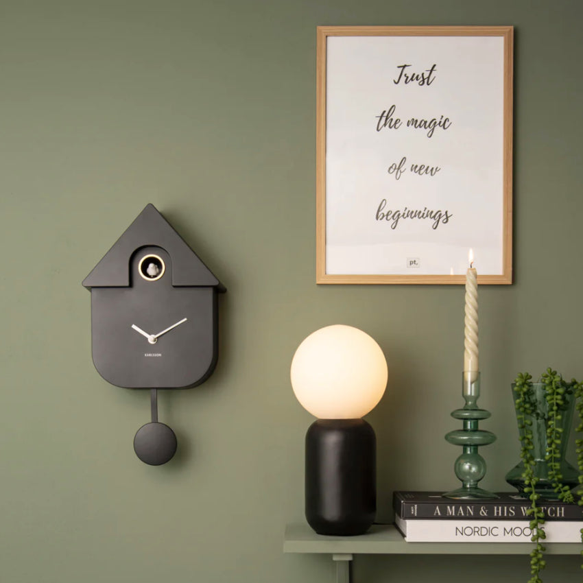 The Karlsson Modern Cuckoo Clock in Black on a green wall next to a shelf