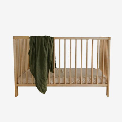 The Leni The Label Muslin Swaddle in Olive hanging over a crib on a blank background