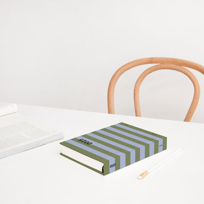 The Lettuce Daily Diary in Periwinkle Olive Stripe on a table with a magazine and pencil next to it