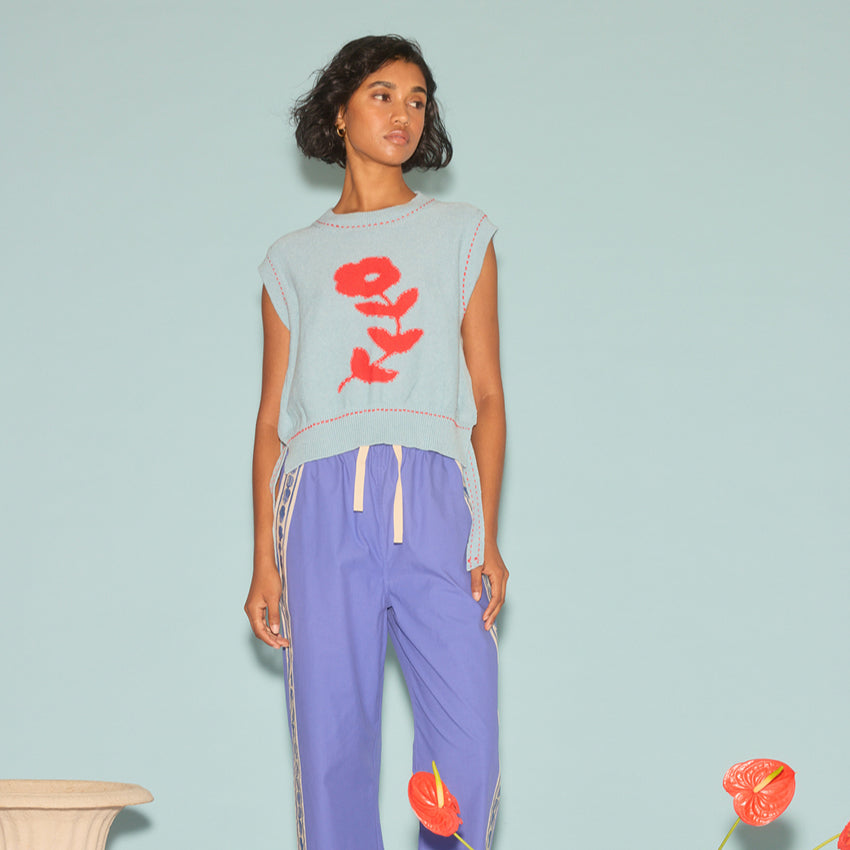 The Nancybird Aria Vest in Flower on a model paired with track pants on a blue background