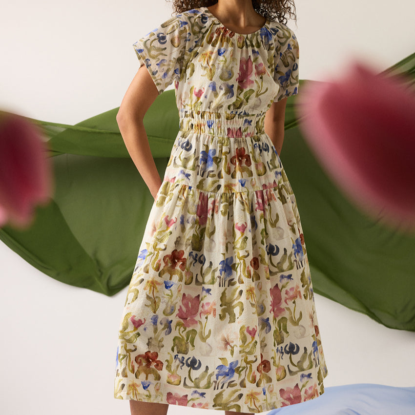 A model wearing the Nancybird Lola Dress in Watercolour Collage with fabric and flowers around her.