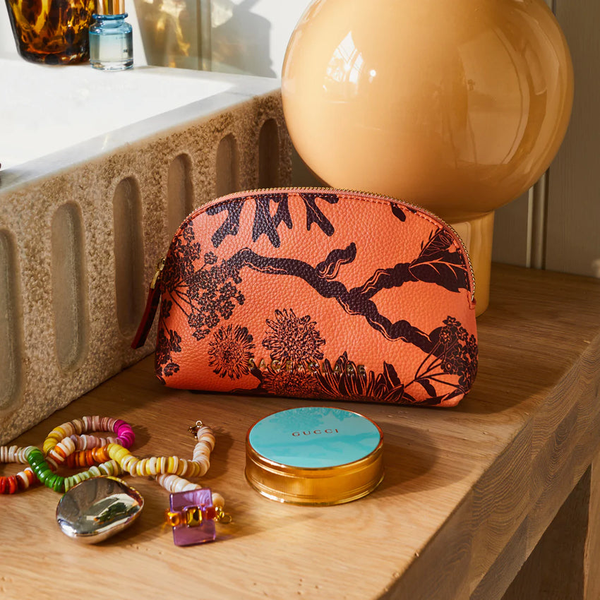 The Sage x Clare Delphine Cosmetic Pouch in Vivid Coral on a vanity with makeup and jewellery next to it