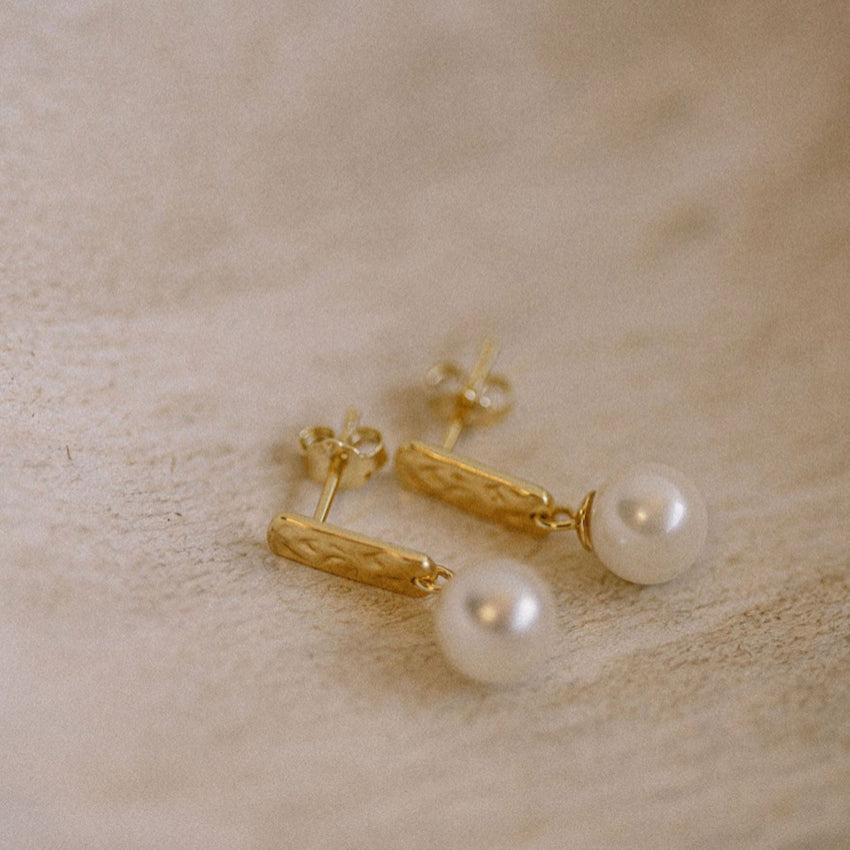 The Silver Linings Collective Luna Pearl Earrings in Gold on a textured background