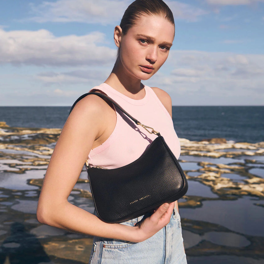 The Status Anxiety Look Both Ways in Black and Brush Gold on a models shoulder with the short strap in front of the sea