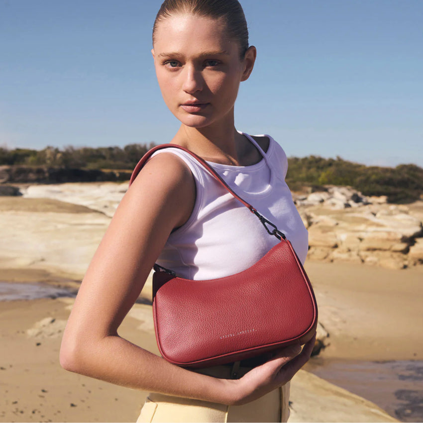 A model wearing the Status Anxiety Look Both Ways Bag in Rouge on the shoulder with the short strap