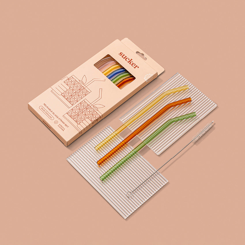 The Sucker Reusable Glass Straws in Multi in the packaging and next to 3 of the staws and cleaner