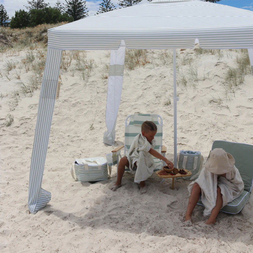 A kid sitting on the SunnyLife Deluxe Beach Chair in Sea Sage Stripe on the beach under a pop up 