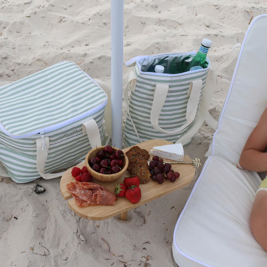 The SunnyLife Light Cooler Bag in Sea Sage on the beach next another cooler bag and cheese board