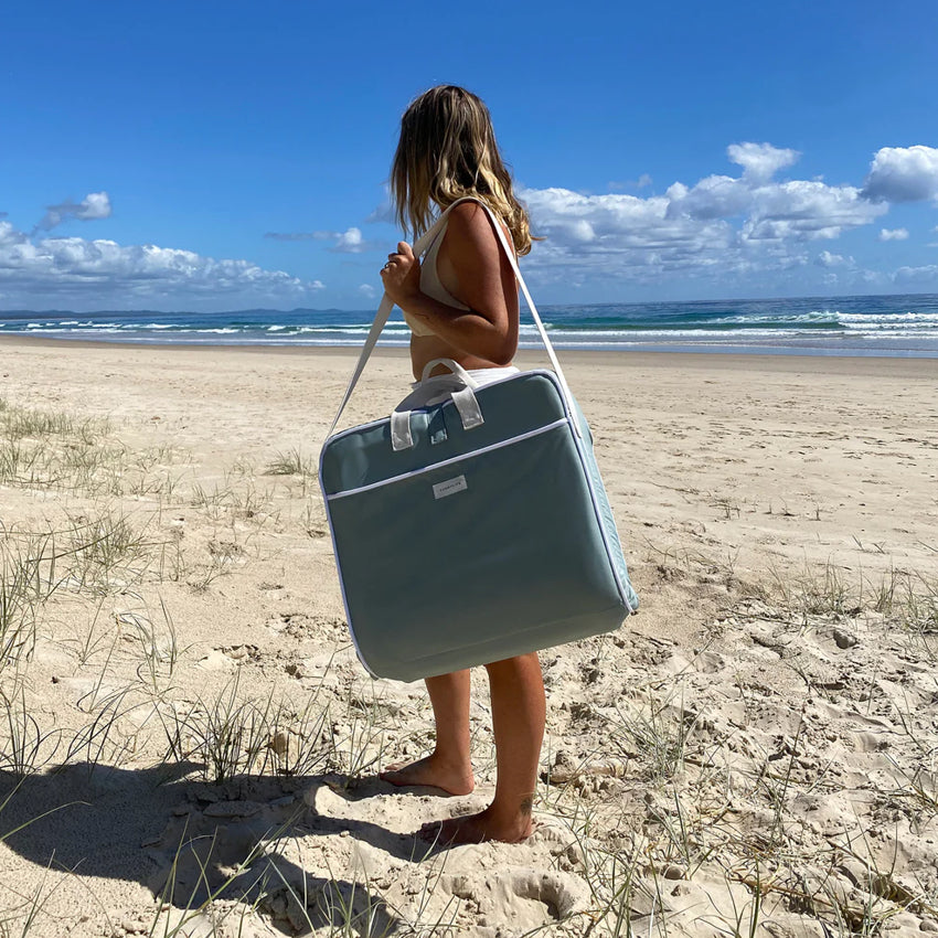 A woman holding the Sunny Life Take Anywhere Folding Chair in Sea Sage folded up on her shoulder on the beach
