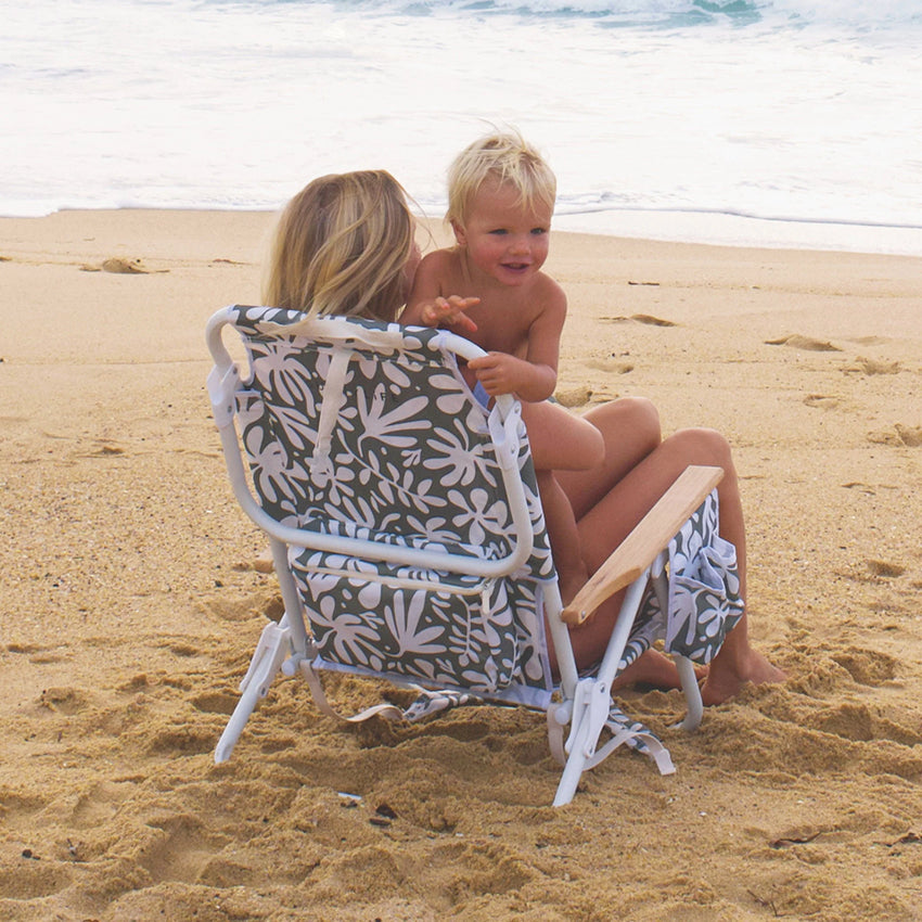 The Sunny Life Luxe Beach Chair in Olive on the beach with a lady and child sitting in it