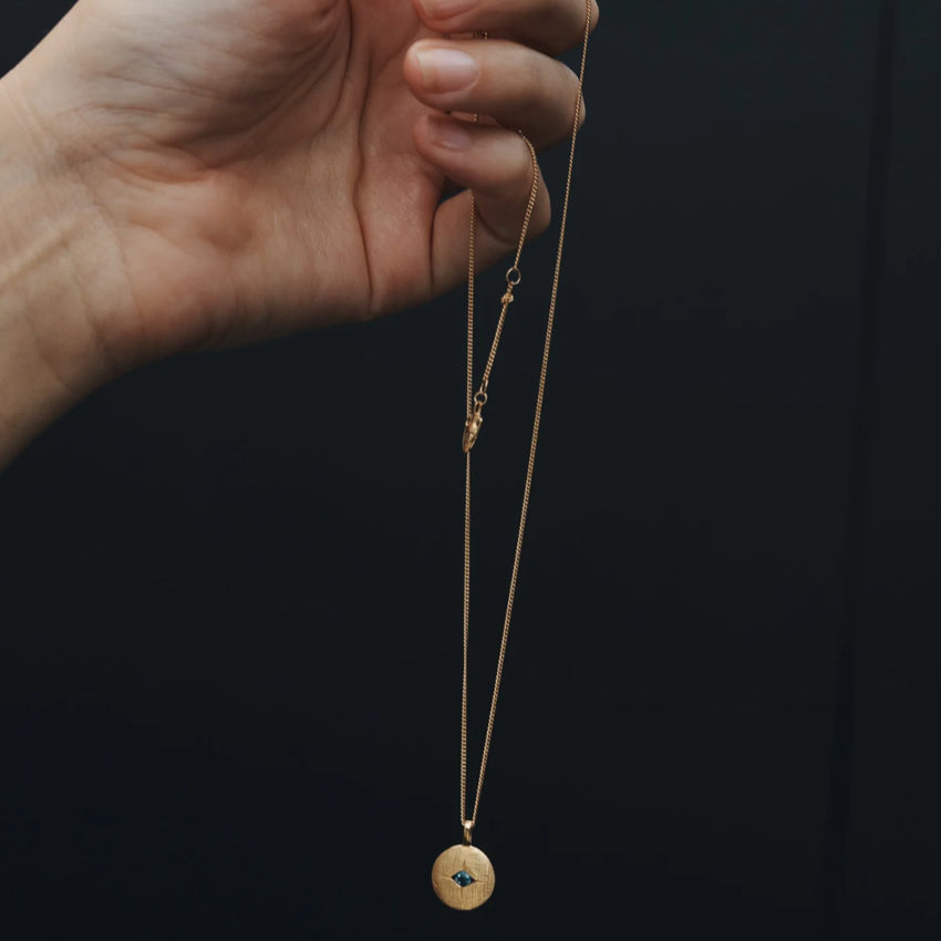 The Temple Of The Sun Mavi Eye Necklace in Gold being held in front of a black background