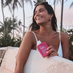 A model holding the We Are Feel Good Inc Wild Rosella SPF 50+ Sunscreen Lotion 200ml with a surfboard