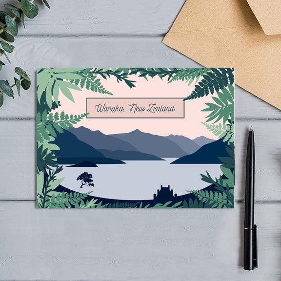 Greeting card with a scenic design of mountains and lake, featuring the text 'Wanaka, New Zealand', on a wooden surface with a pen and envelope.