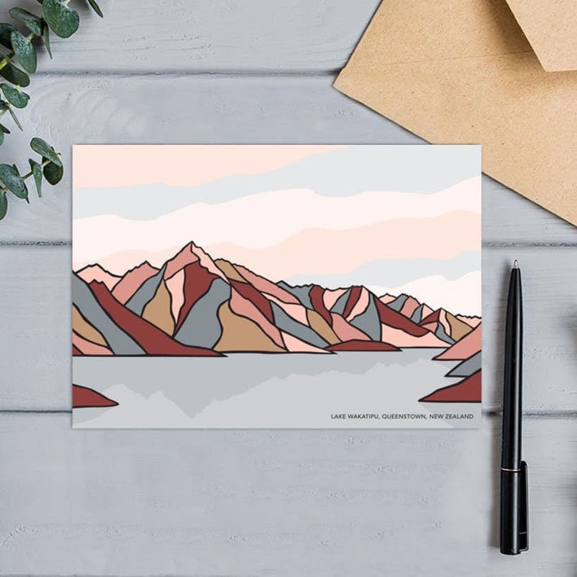 A greeting card with a modern landscape design of Lake Wakatipu, Queenstown, on wood next to a pen and envelope