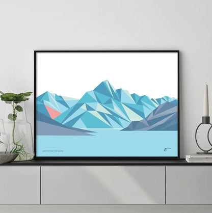 A geometric art print of Mt Cook in blue and pink hues, by Bridget Hall in a black frame on a console.