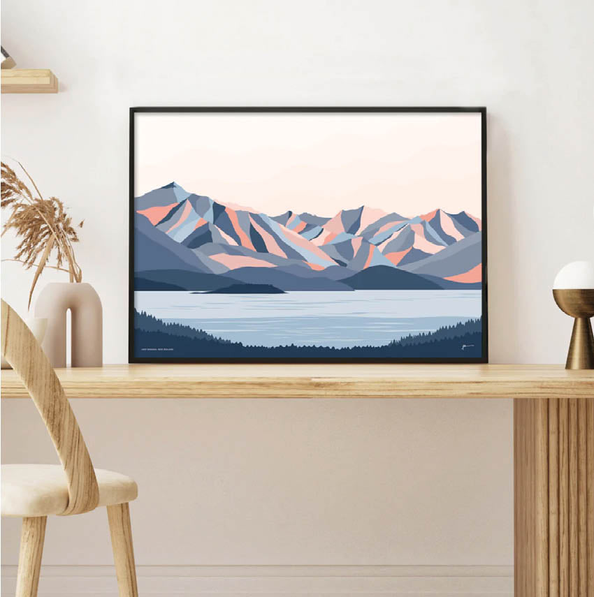 A contemporary art print of the mountains surrounding Wánaka, New Zealand by Bridget Hall Designs in a black frame on a desk.