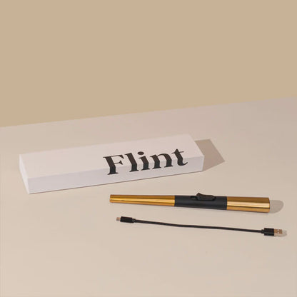 The Flint Rechargeable Lighter in Gold next to its box and charger